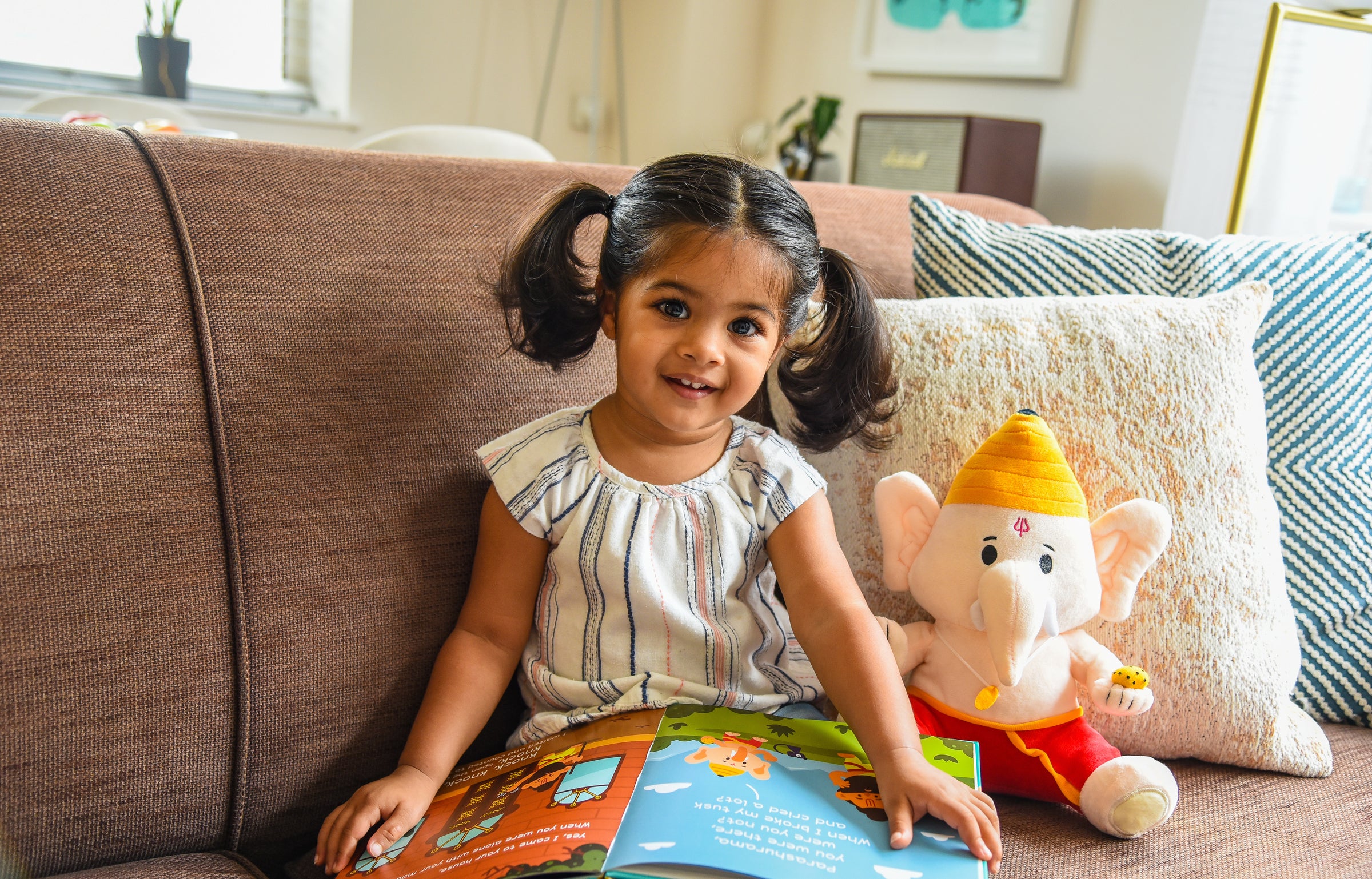 child next to ganesh plush toy on couch reading modi toys book
